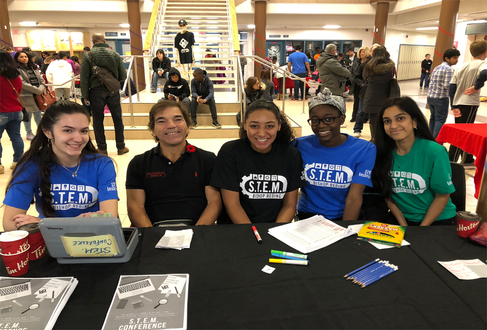 People watching towards camera and smiling,table with book and pens on it,back ground have stairs and more peole